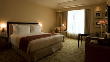 A hotel room with a large bed, two bedside tables with lamps, a desk with a chair, and a large window with curtains.