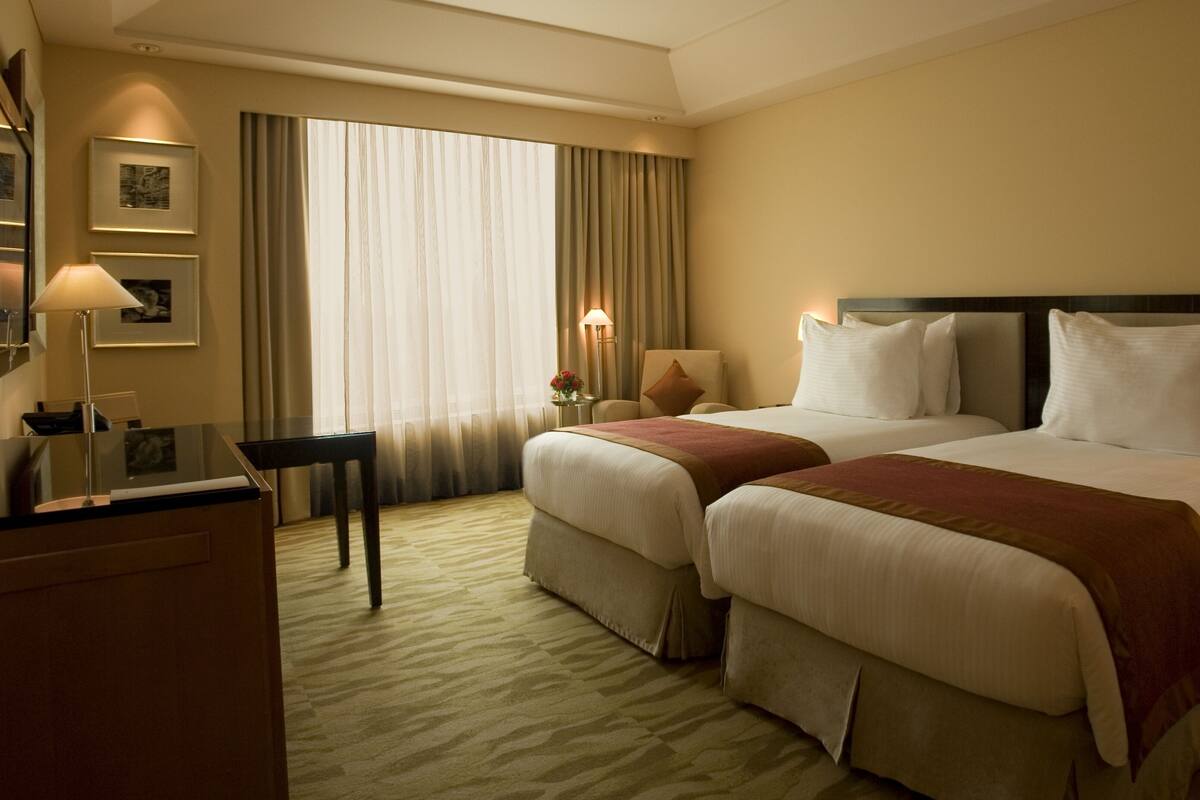 A hotel room with two beds, a desk, a chair, and a large window with curtains.