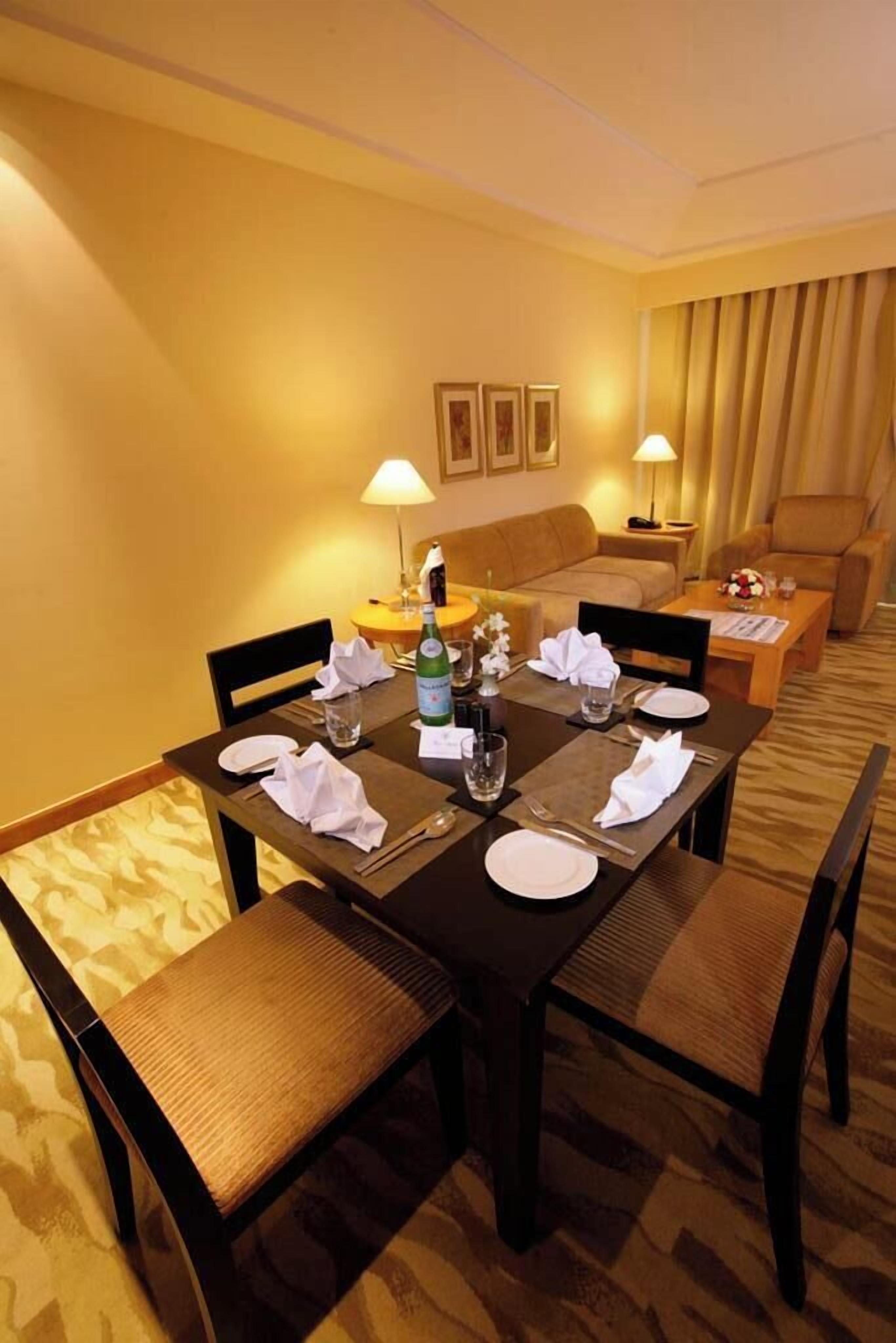 A hotel dining area with a rectangular table set for four, surrounded by cushioned chairs. There are two wall-mounted lamps, a sofa, and a small side table with a vase of flowers.