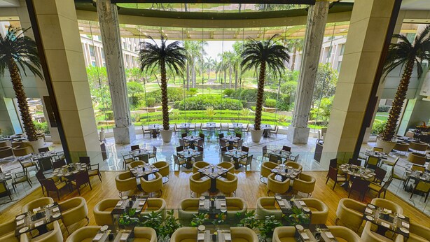 Un'ampia sala da pranzo interna con numerosi tavoli e sedie, una piscina centrale e vista su un giardino curato con palme.