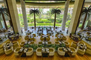 A spacious indoor dining area with multiple tables and chairs, a central pool, and a view of a well-maintained garden with palm trees.