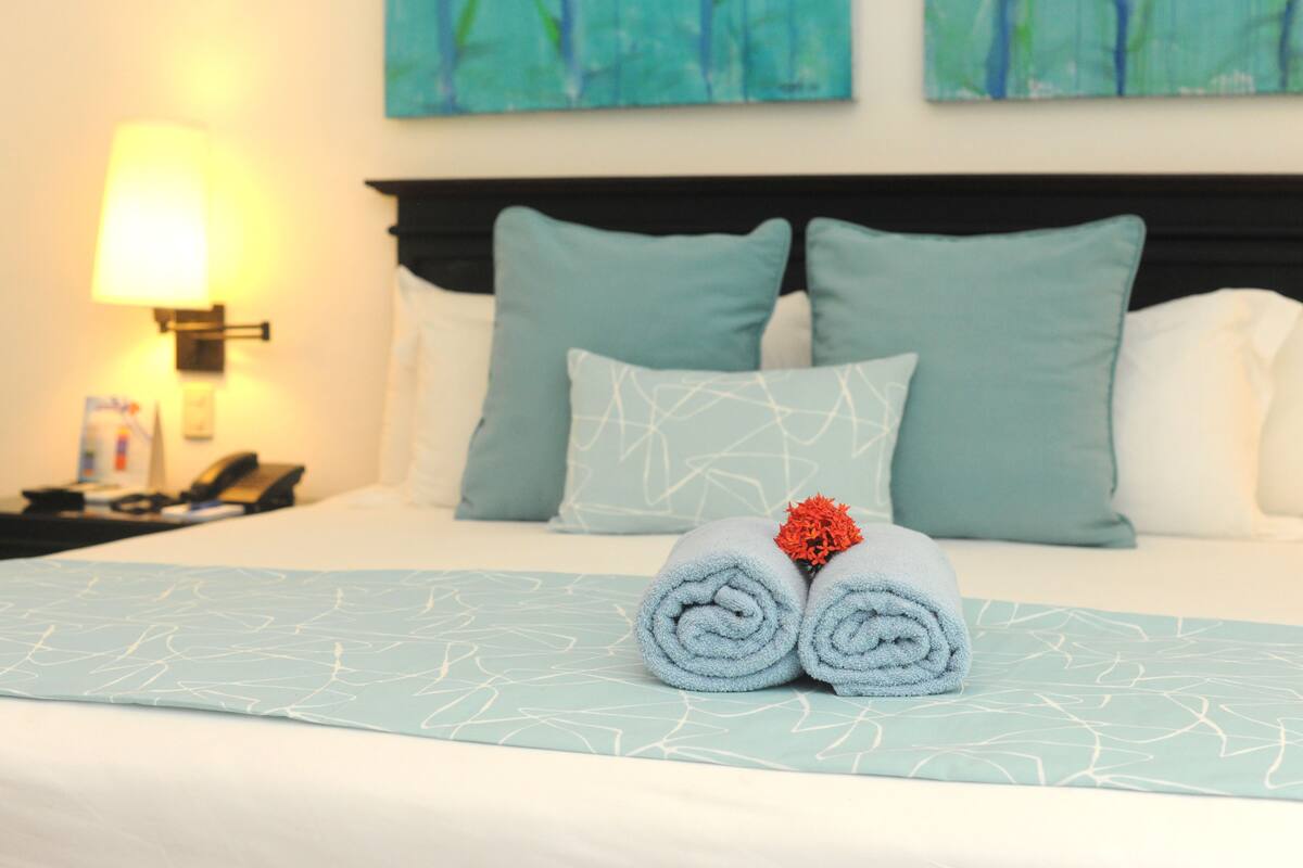 A neatly made bed with a blue and white patterned bedspread, two rolled towels with a red flower, and matching pillows.