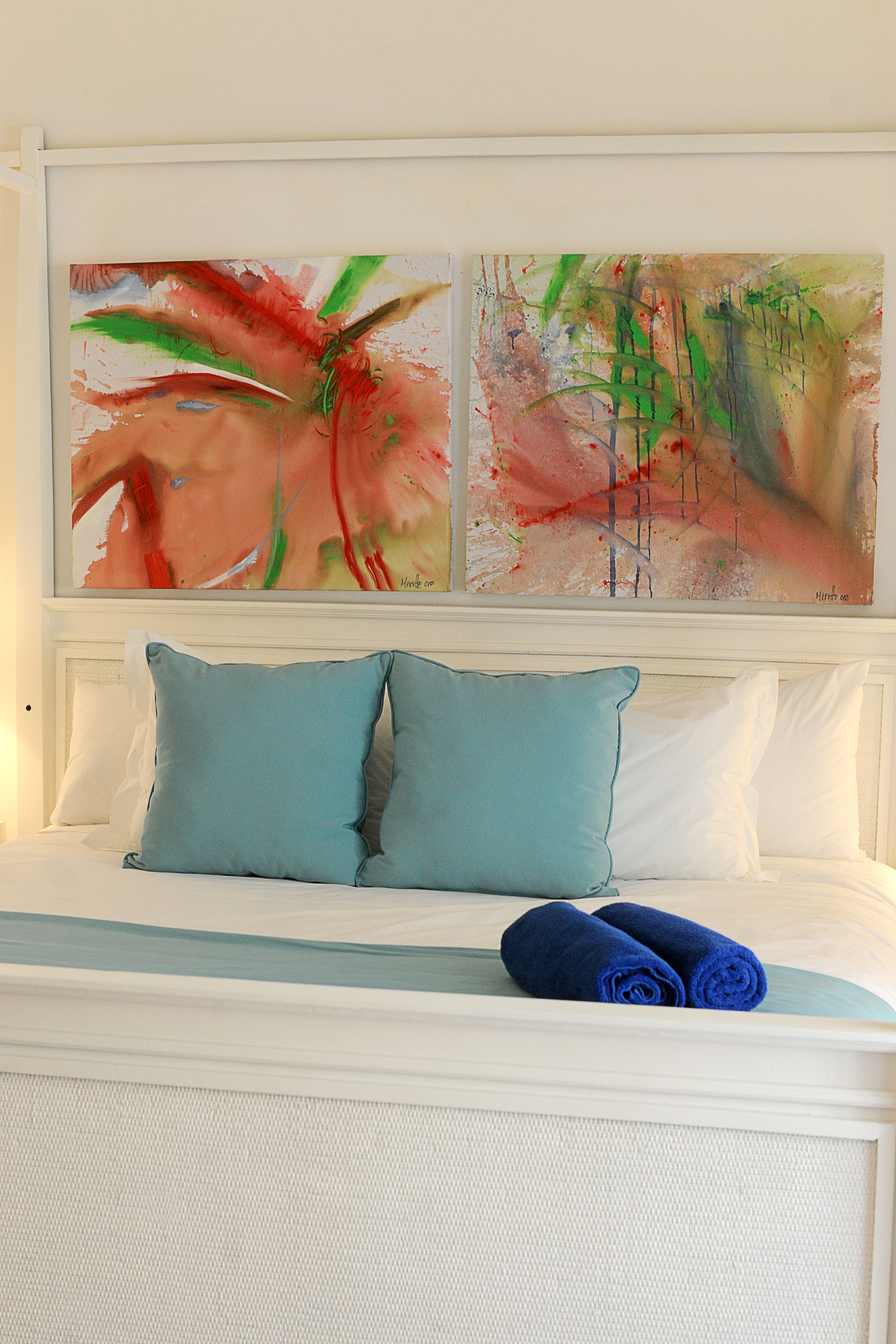 A bed with two pillows, a blue towel, and two abstract paintings above.