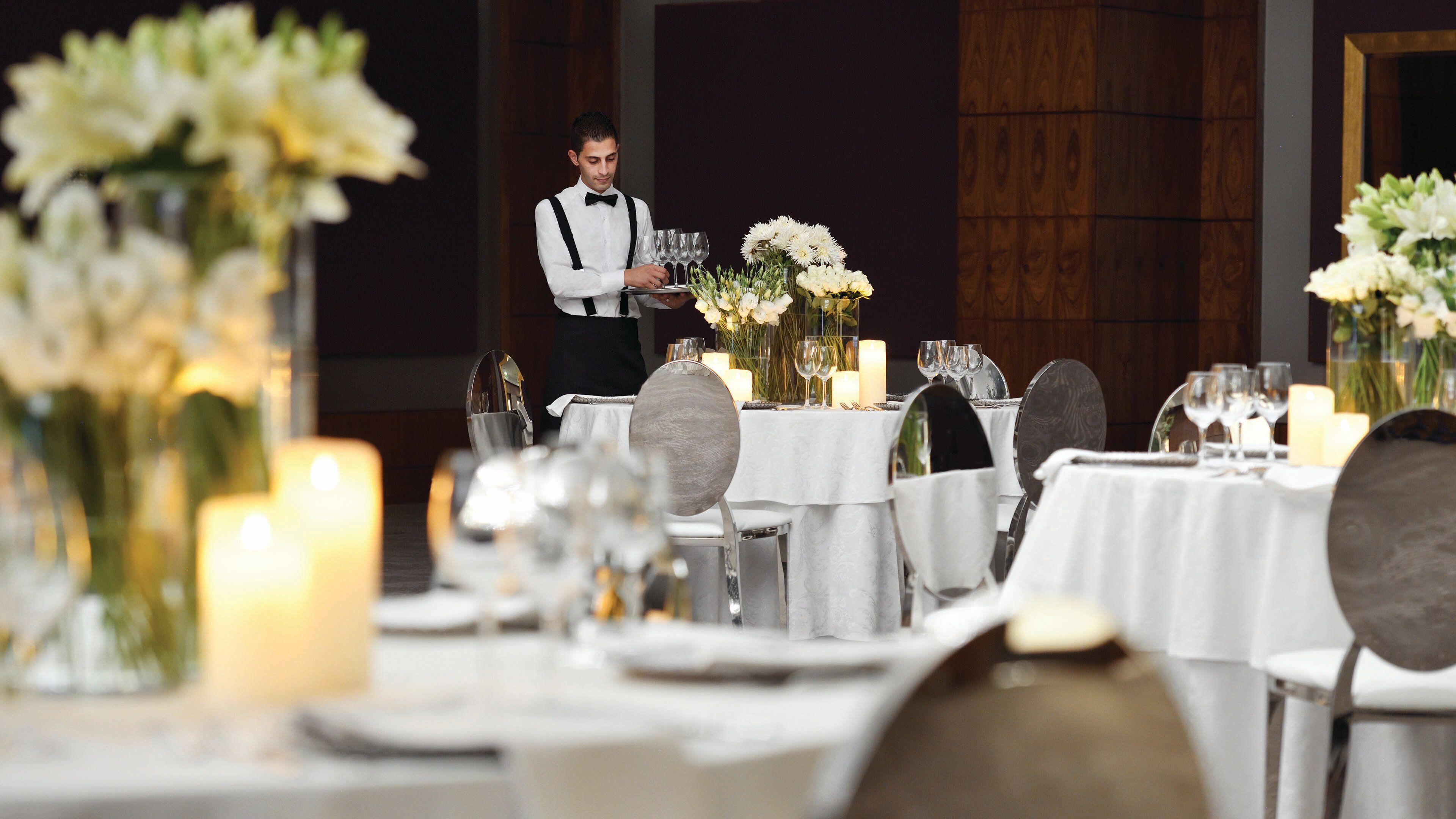 A formal dining setup with tables set for guests, a waiter attending to the table, and floral centerpieces.