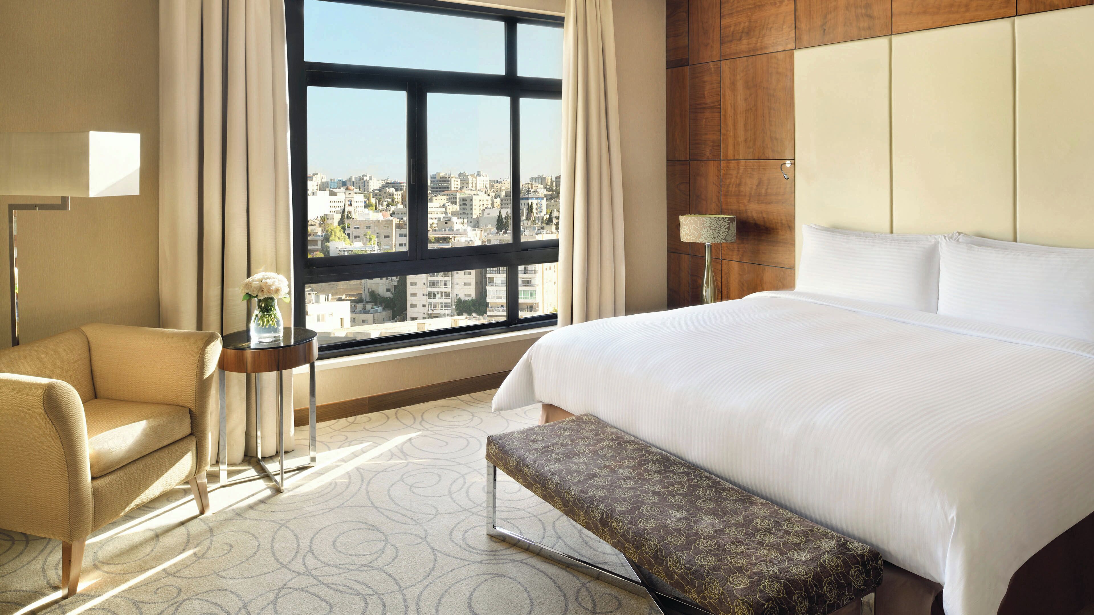 A hotel room with a large bed, a bench, a chair, a small table with a vase, and a view of the city through a large window.