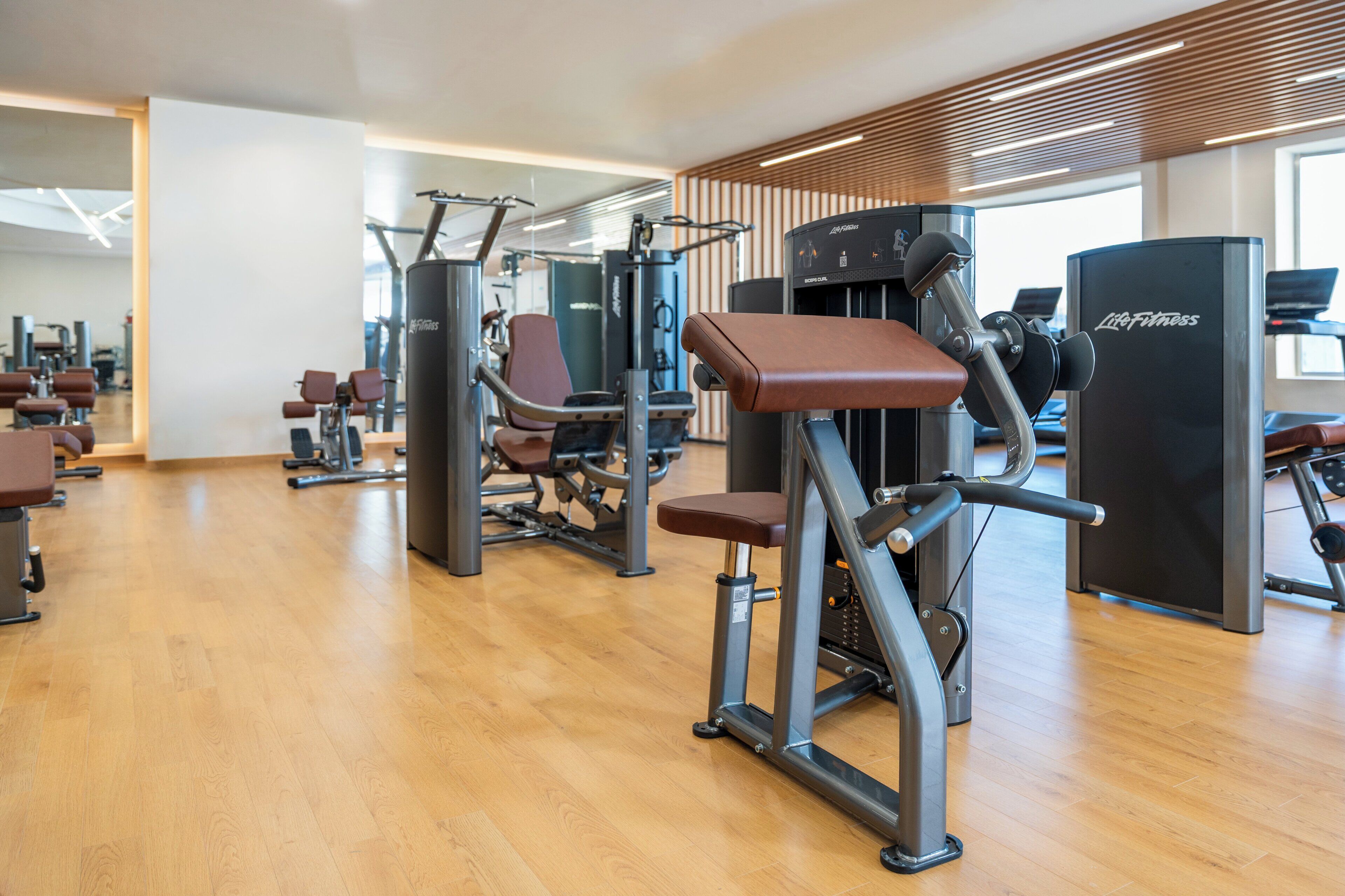 A gym with various exercise machines, wooden flooring, and large windows allowing natural light.
