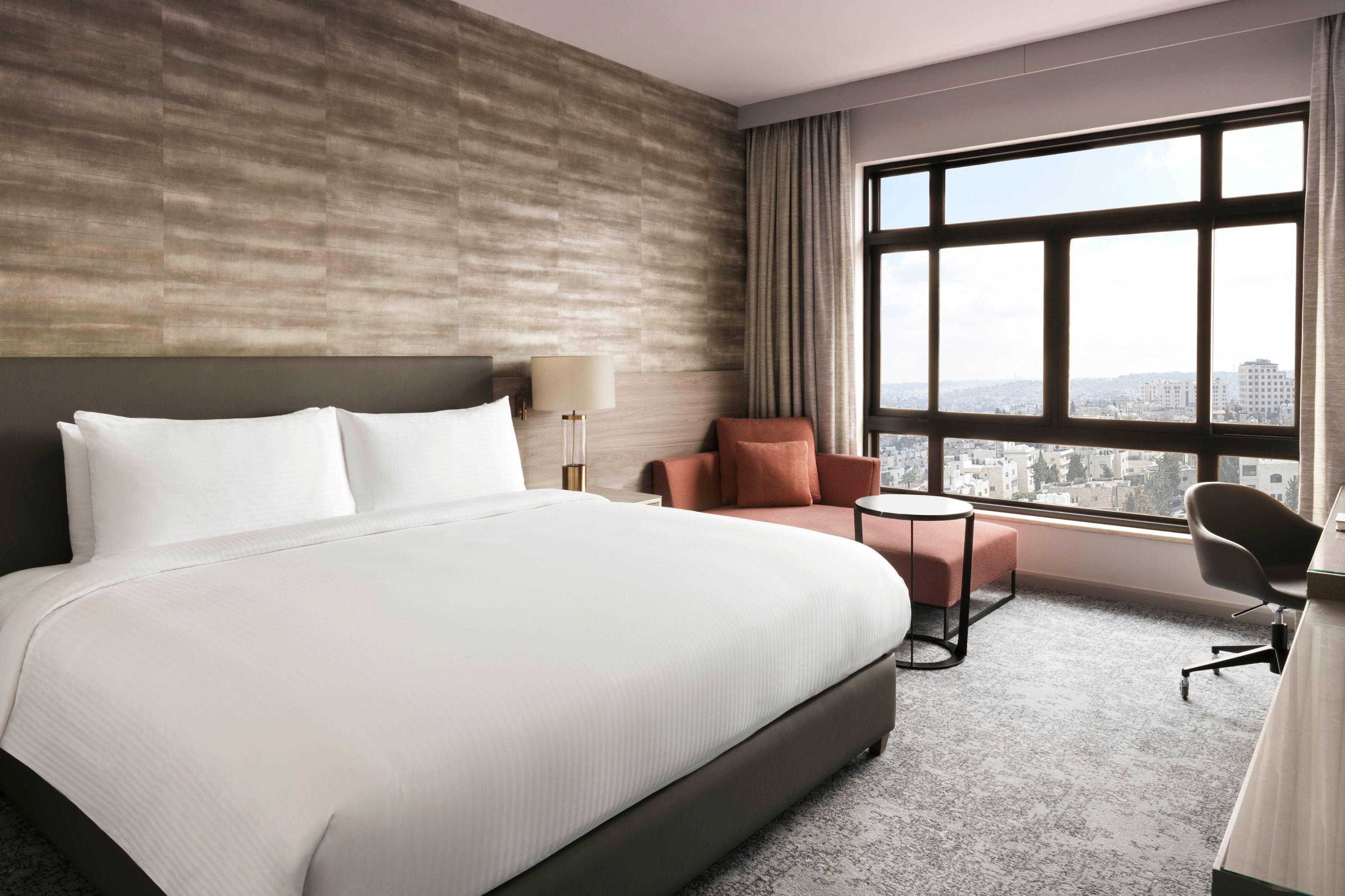 Une chambre d’hôtel avec un grand lit, un bureau avec une chaise, une petite table et une vue sur la ville par une grande fenêtre.