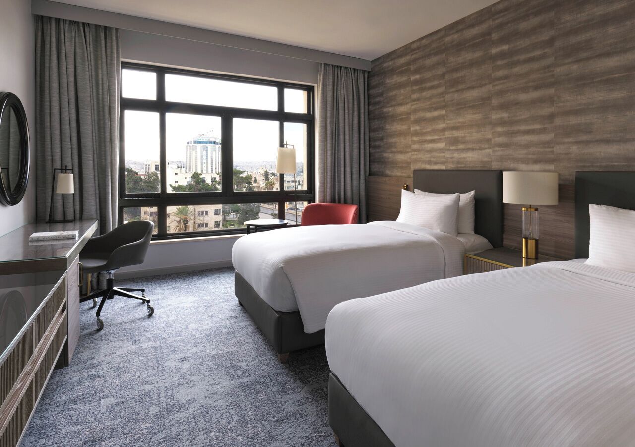 Une chambre d’hôtel avec deux lits, un bureau, une chaise et une grande fenêtre donnant sur la ville.