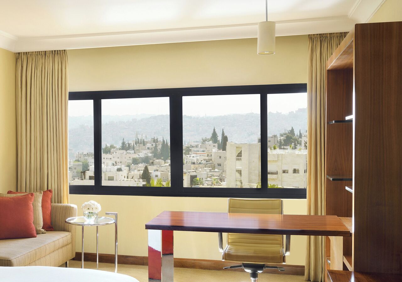 A hotel room with a large window, a sofa, a small round table, and a wooden desk.