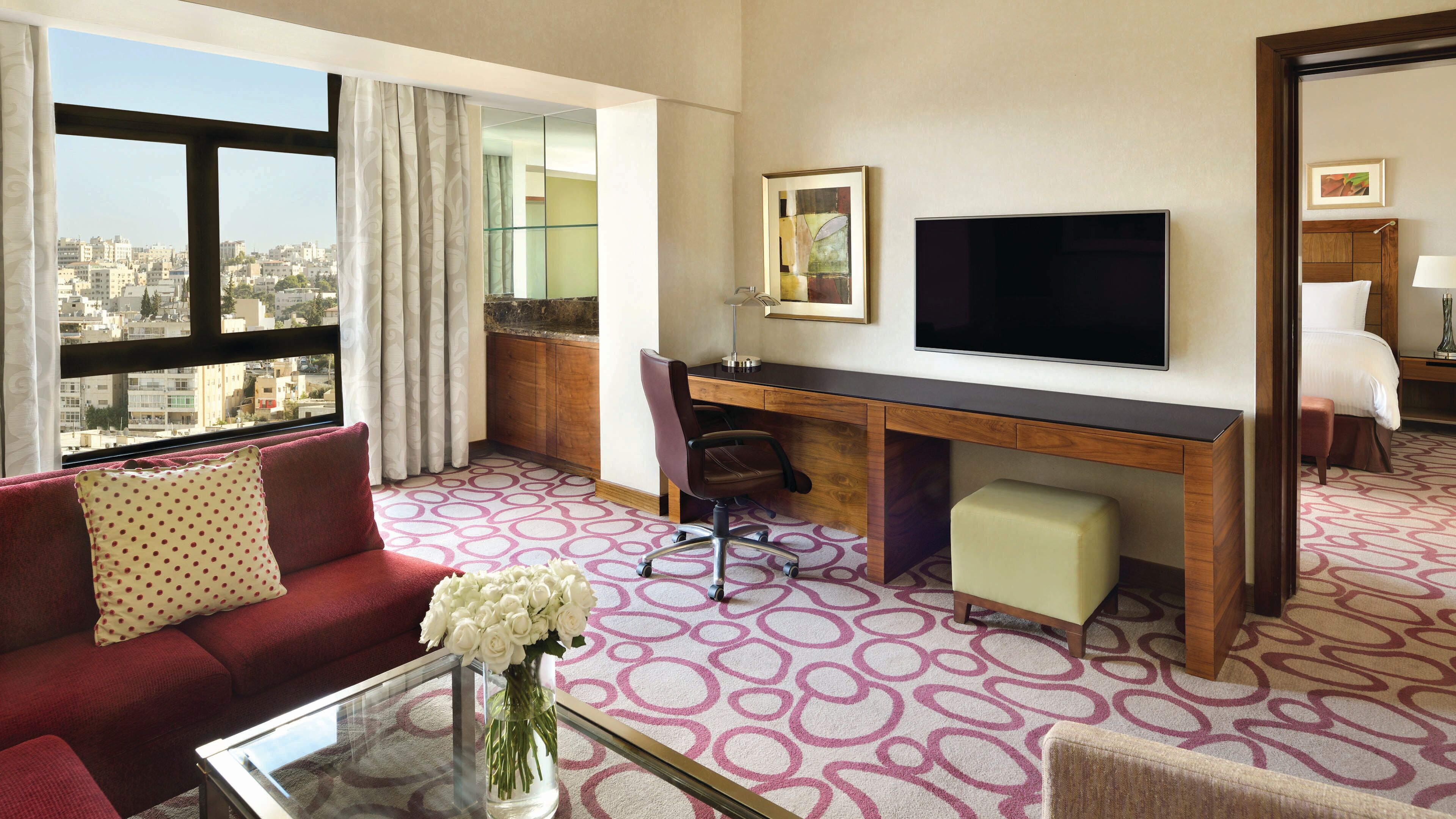 A hotel room with a large window, a desk, a TV, a sofa, and a bed.