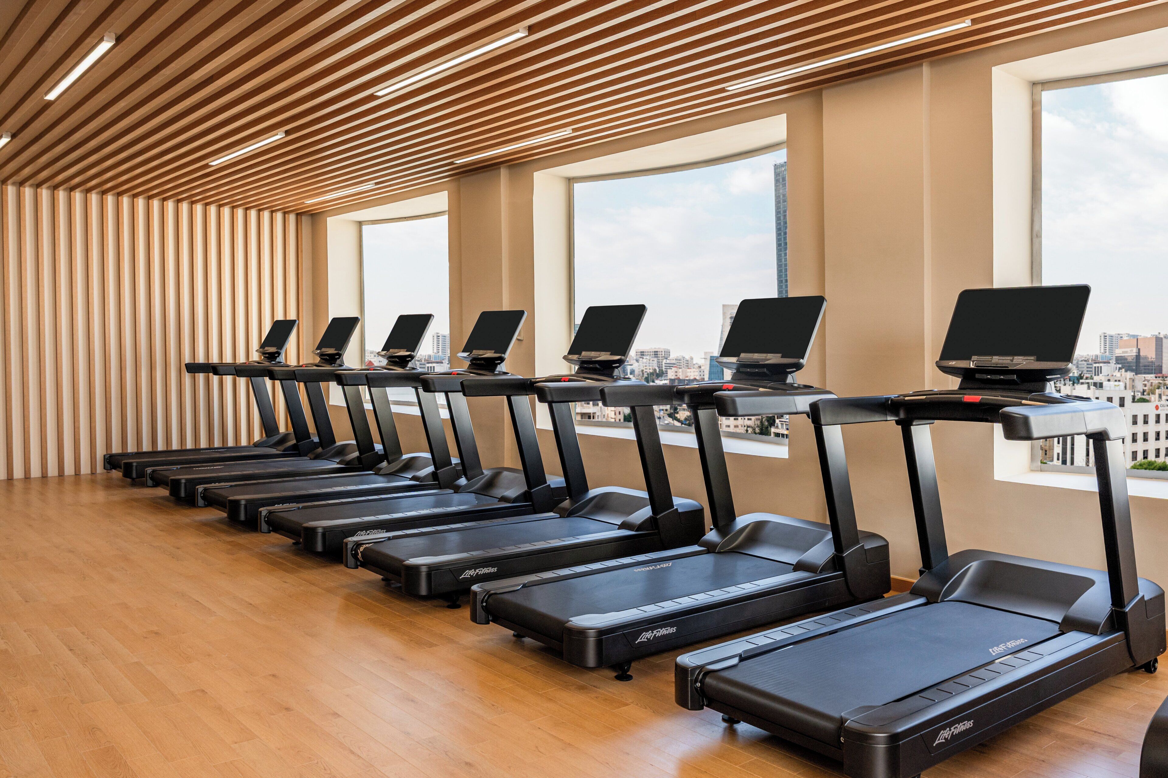 A row of treadmills with attached screens in a modern room with wooden slats on the ceiling and large windows.