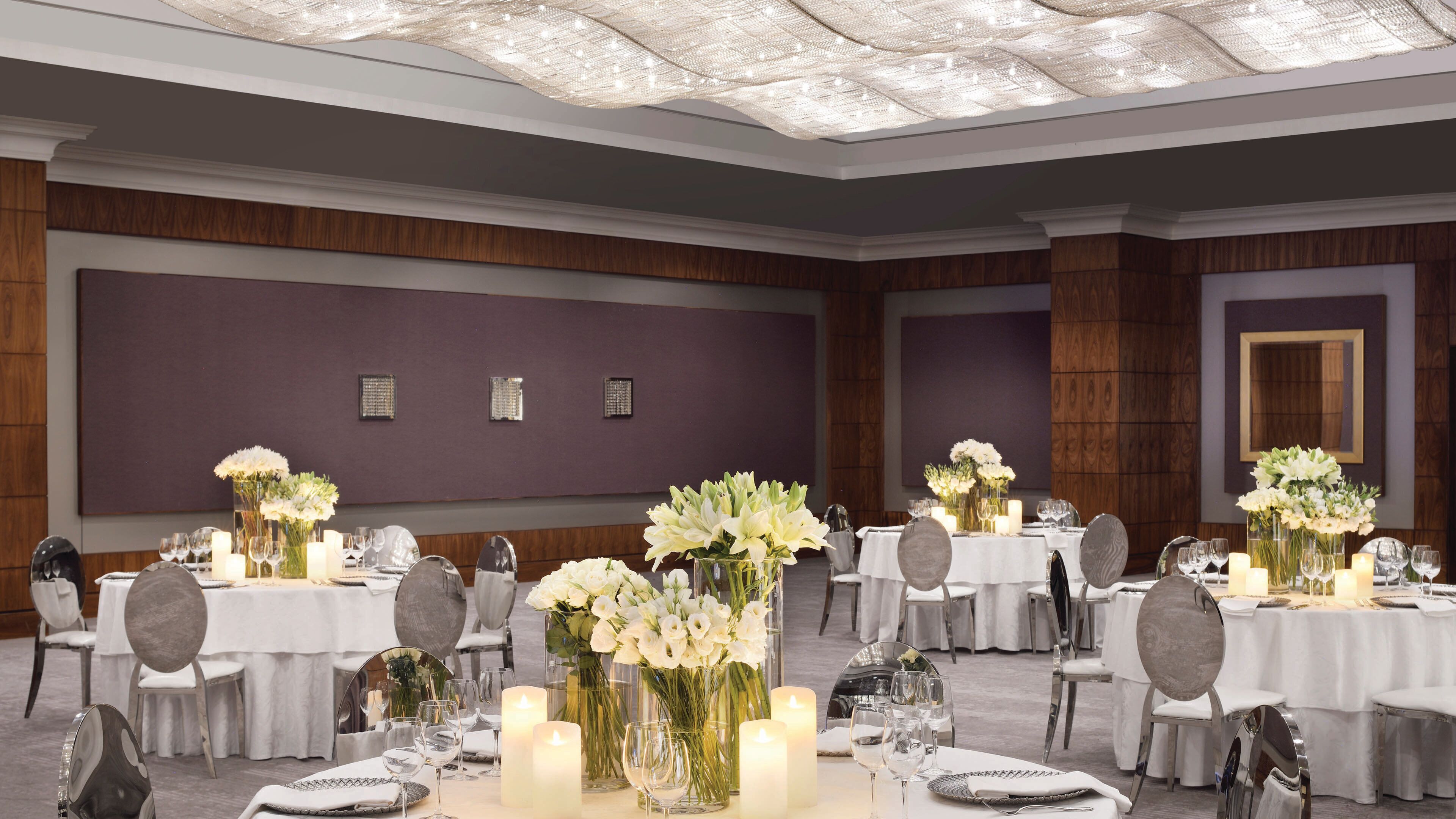 Un salón de banquetes con mesas redondas dispuestas para el servicio de comidas, con manteles blancos, velas y centros de mesa florales.