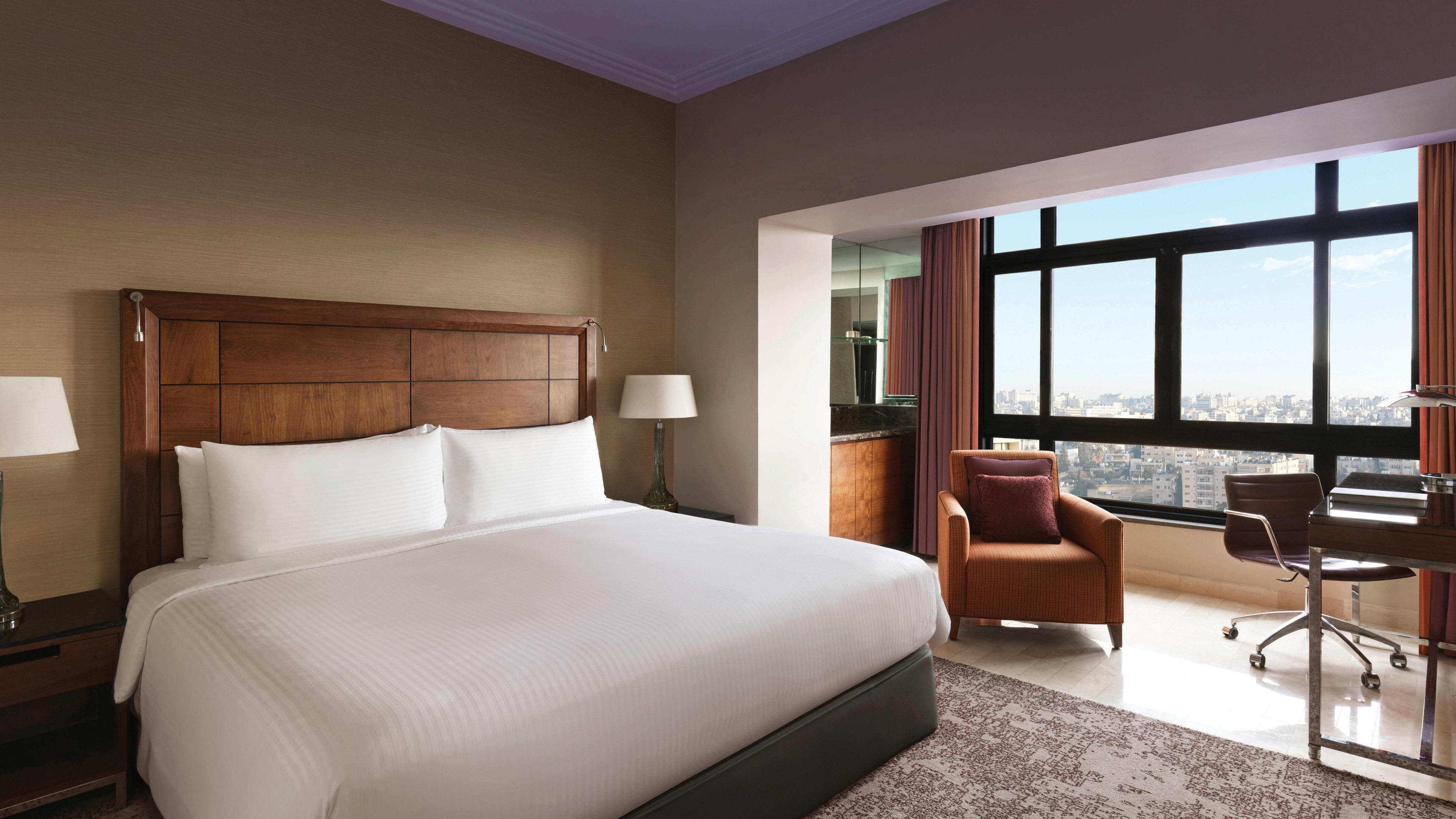 Une chambre d’hôtel avec un grand lit, un bureau, une chaise et une vue sur la ville grâce à de grandes fenêtres.