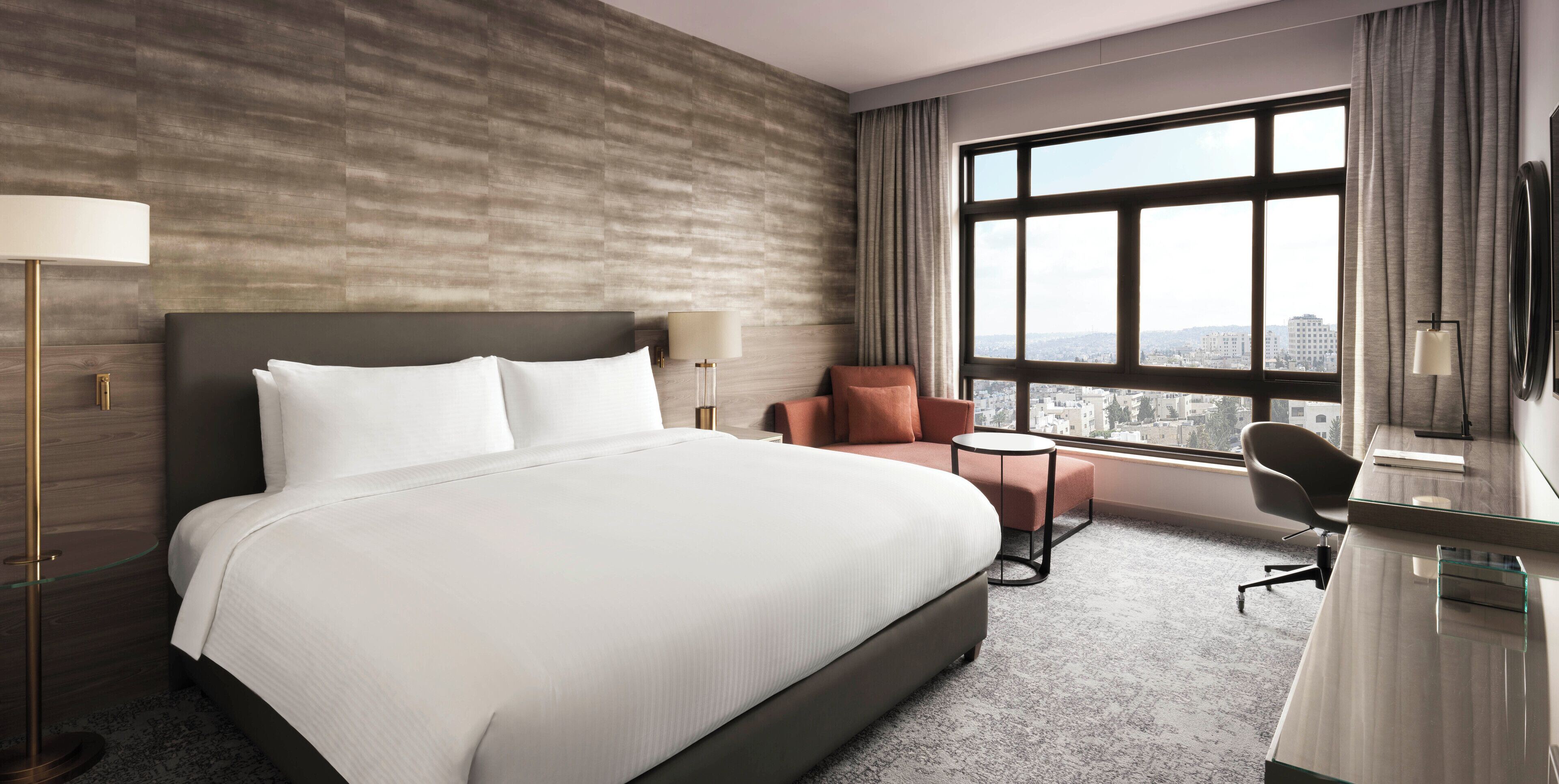A hotel room with a large bed, a desk, a chair, and a view of the city through a large window.