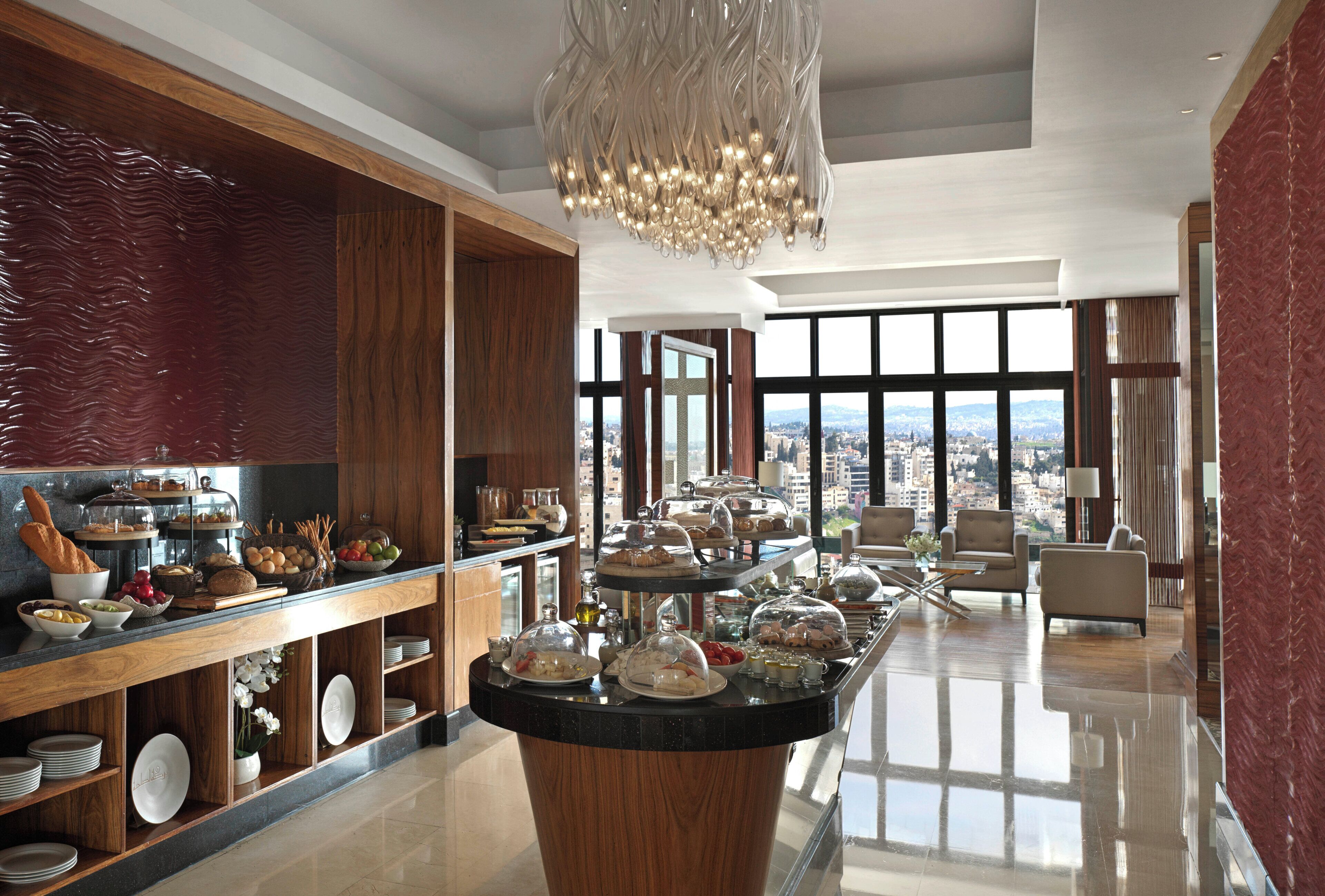 A spacious hotel lobby with a central buffet, large windows offering city views, and a modern chandelier.