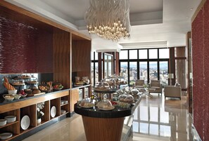 A spacious hotel lobby with a central buffet, large windows offering city views, and a modern chandelier.