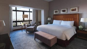 A hotel room with a large bed, a sitting area with a chair and ottoman, a desk, and a view of the city through large windows.
