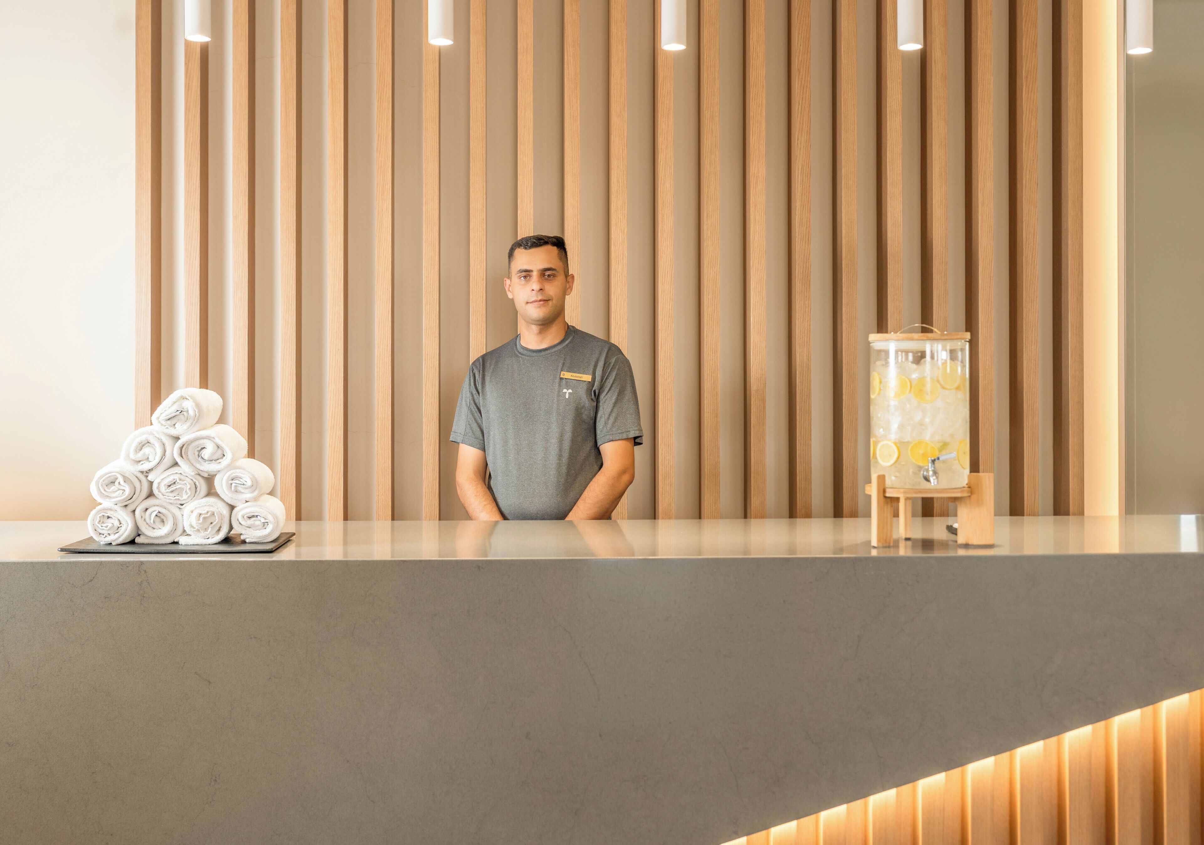 Zona de recepción de un hotel con un panel de madera, mostrador con toallas enrolladas y dispensador de agua.