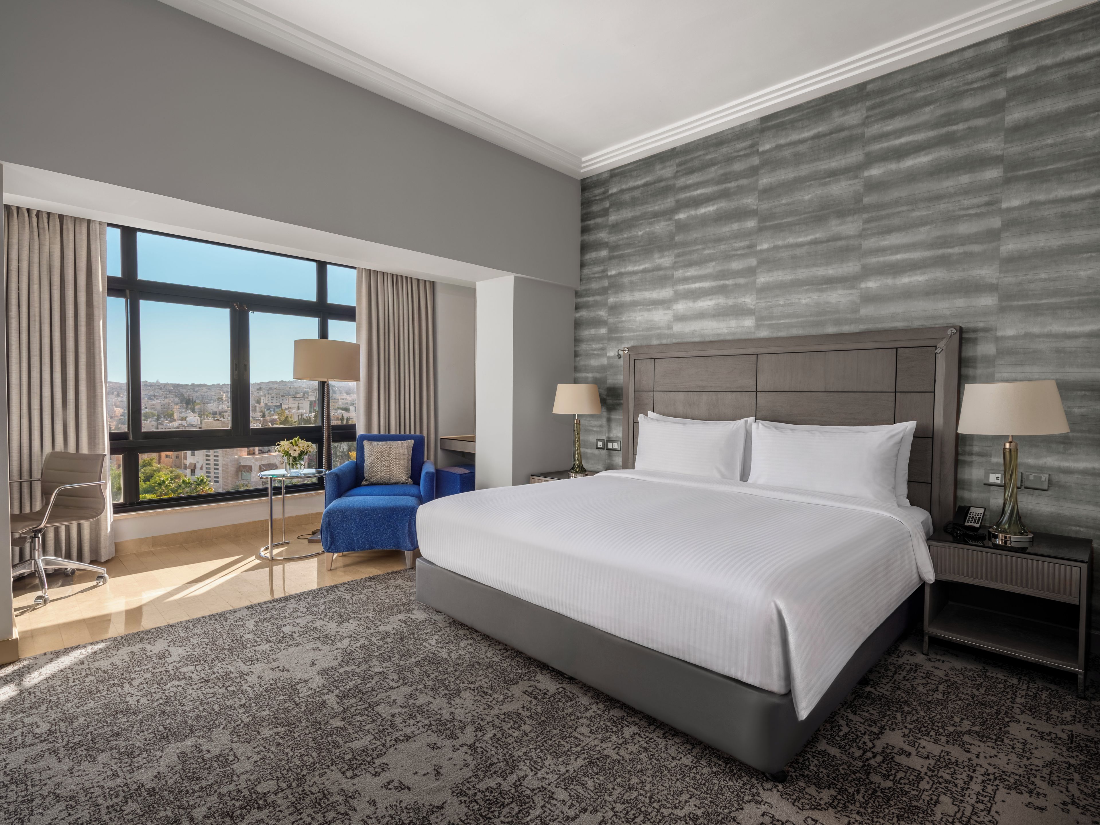 A hotel room with a large bed, a view of the city, a blue armchair, and a desk with a lamp.
