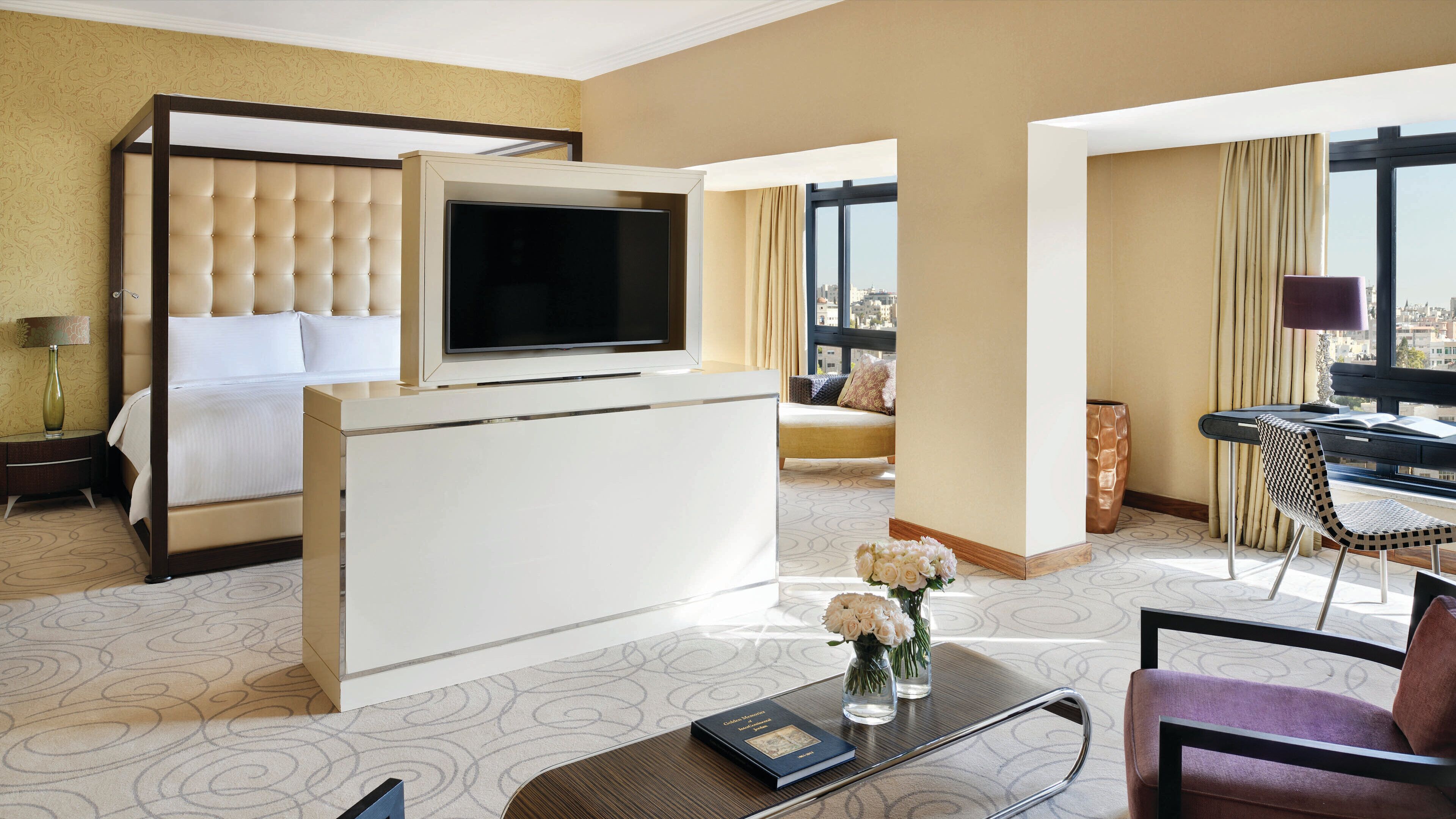 Habitación de hotel con cama, televisor montado en una pared blanca, escritorio con silla y vista a la ciudad a través de amplias ventanas.