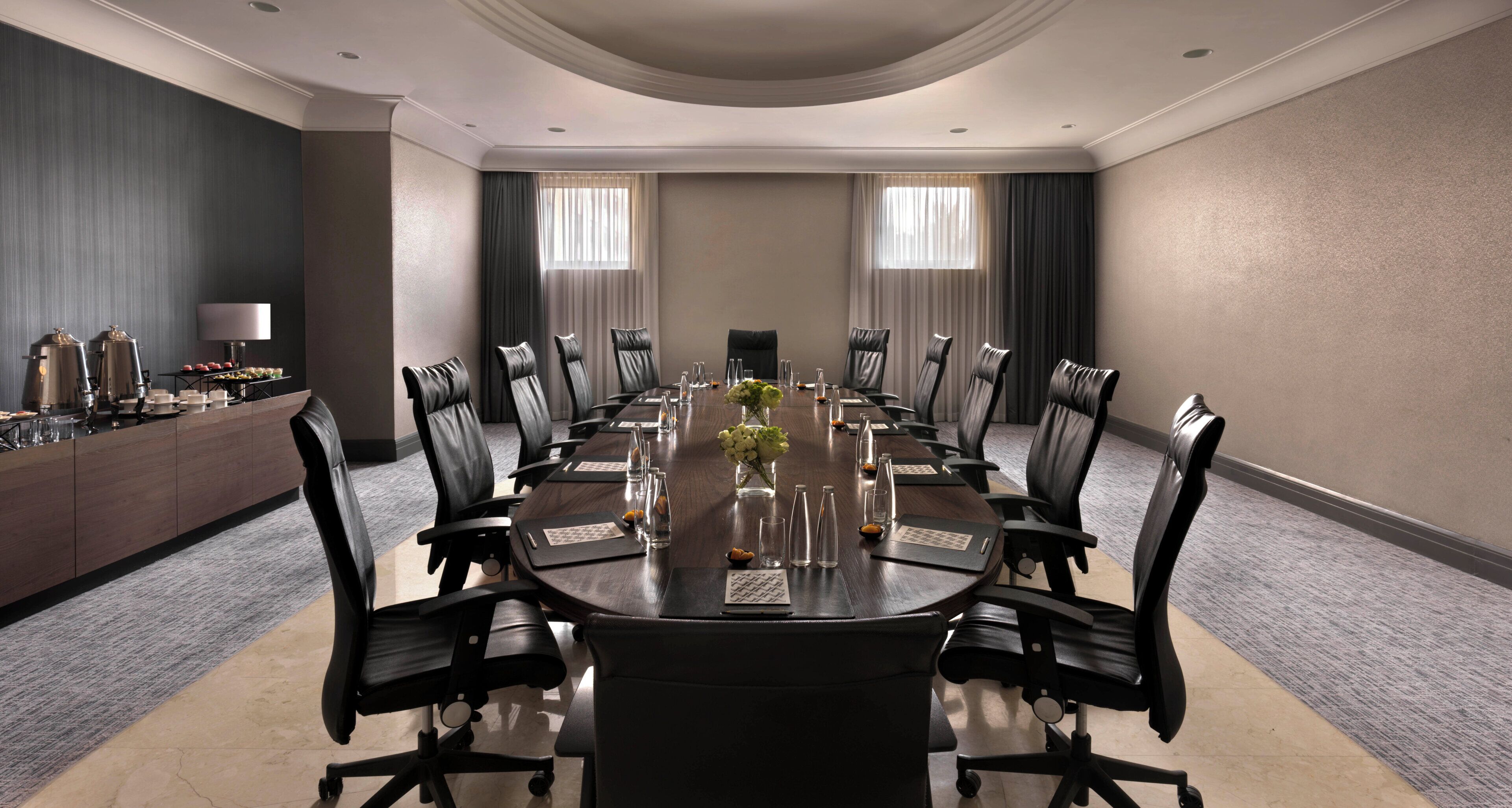 Une salle de conférence avec une longue table, des fauteuils en cuir noir, un coin café et deux fenêtres équipées de rideaux.