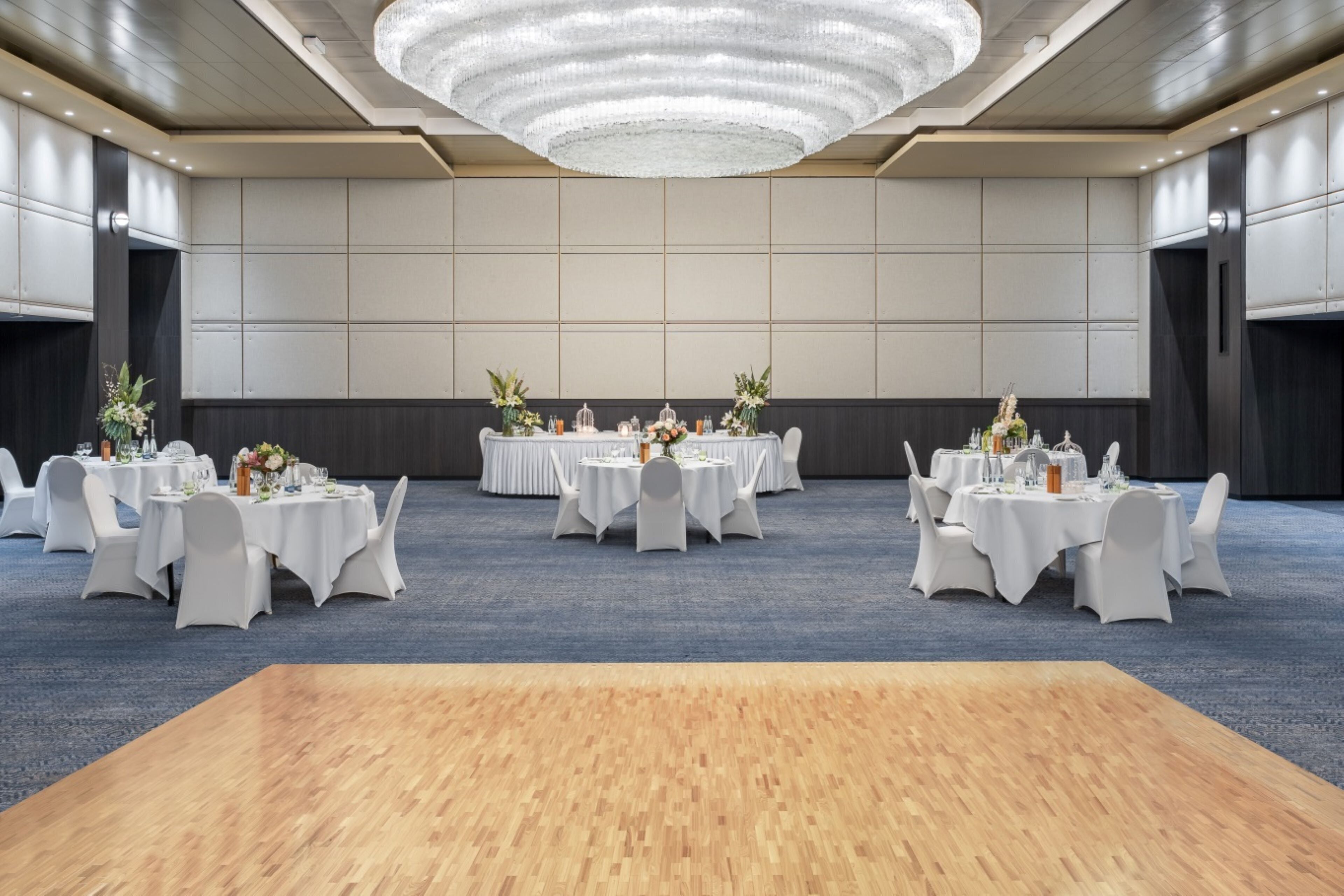 A spacious banquet hall with round tables, white chairs, and a large wooden platform in the center.