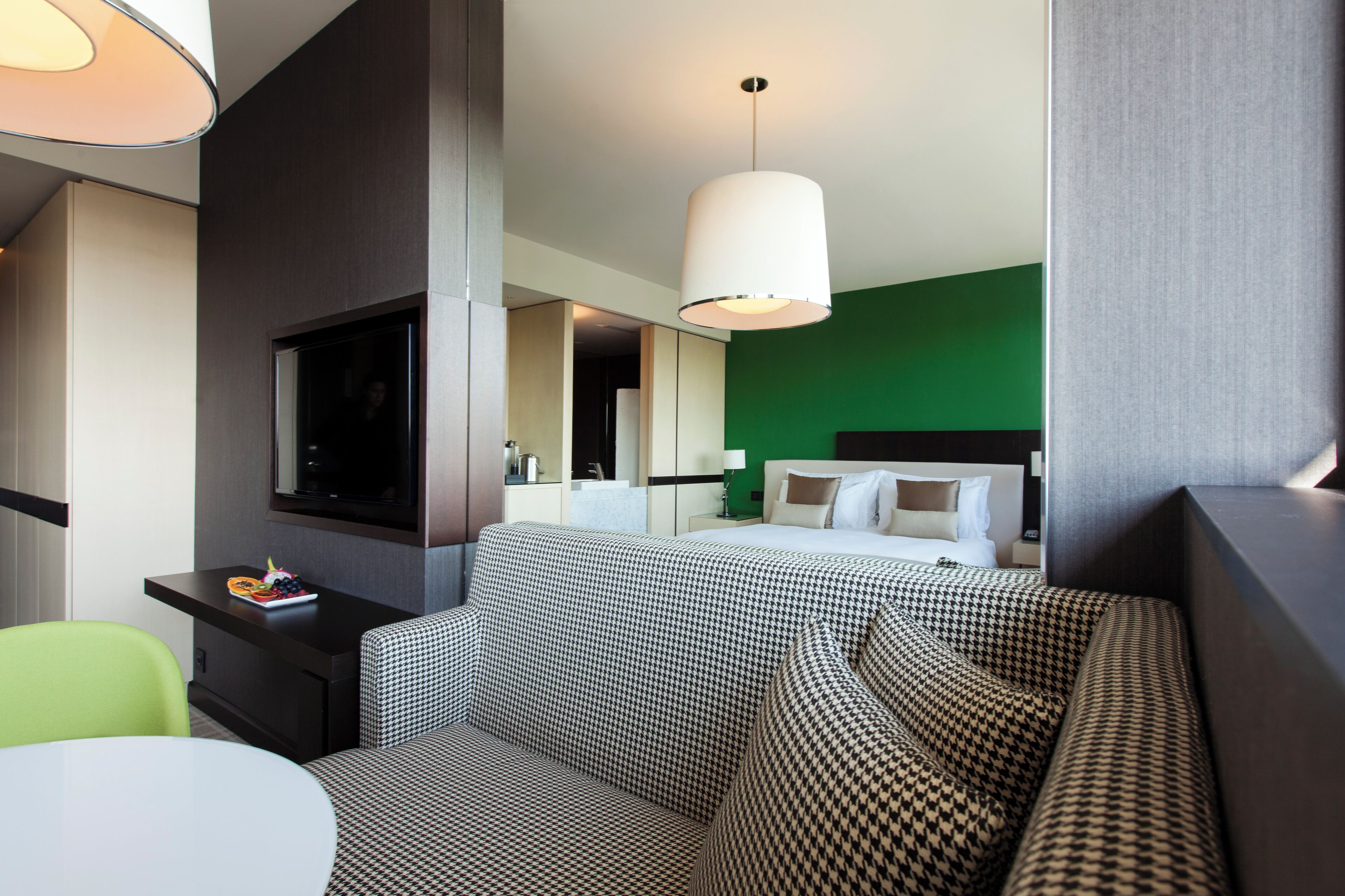 A modern hotel room with a large bed, a sofa, a desk with a TV, and a green accent wall.