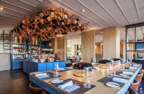 A restaurant with a long dining table set for a meal, a kitchen area with copper pots hanging from the ceiling, and a view of the outdoors through large windows. - Crowne Plaza Geneva by IHG (Meyrin)
