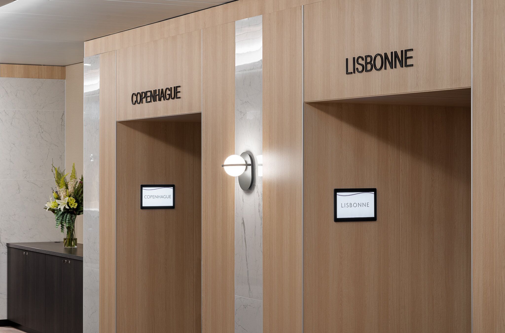 A hotel reception area with wooden paneling and marble accents. There are two signs indicating 'COPENHAGUE' and 'LISBONNE' for room assignments.