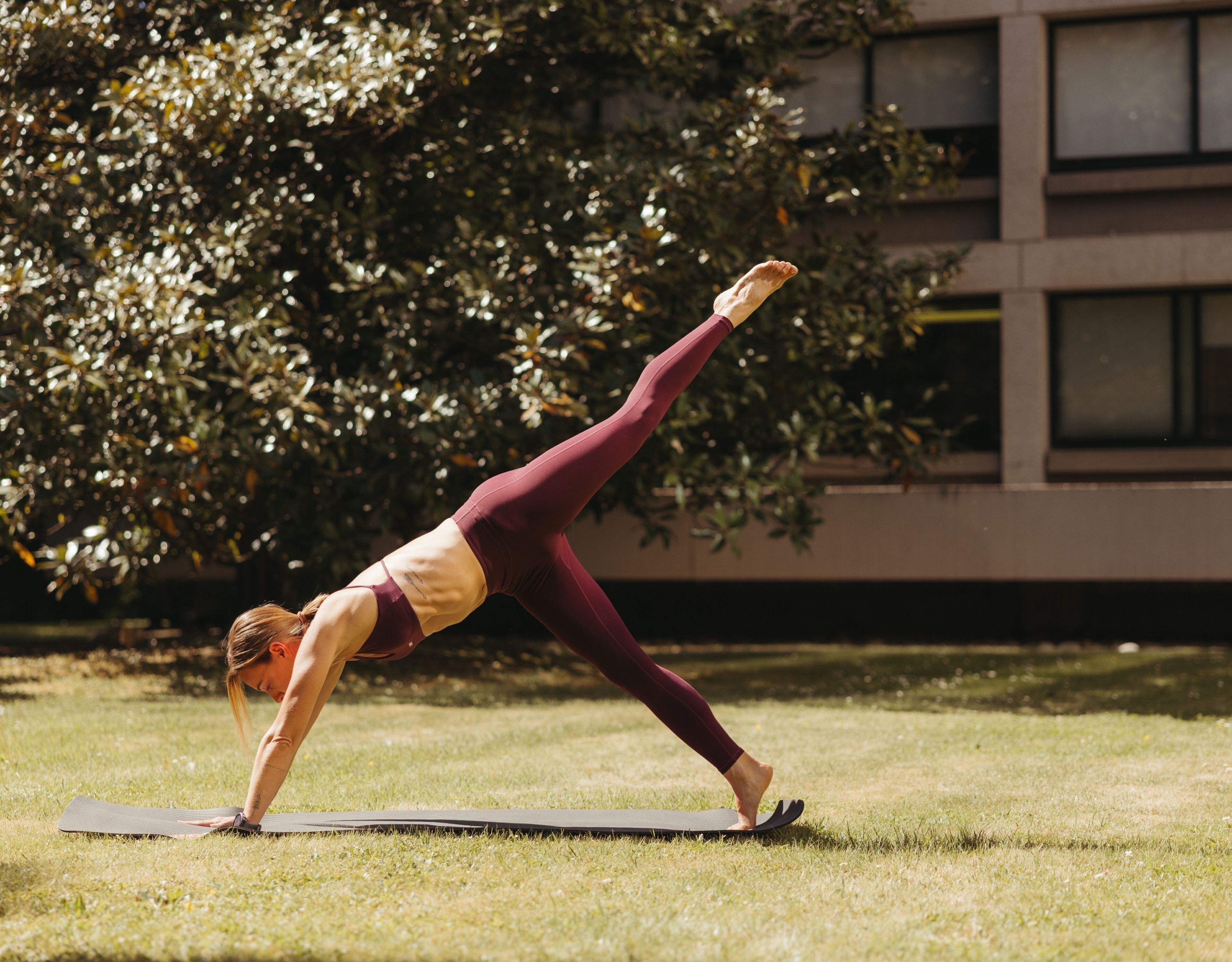 A person performing a yoga pose on a mat outdoors with a building and trees in the background.