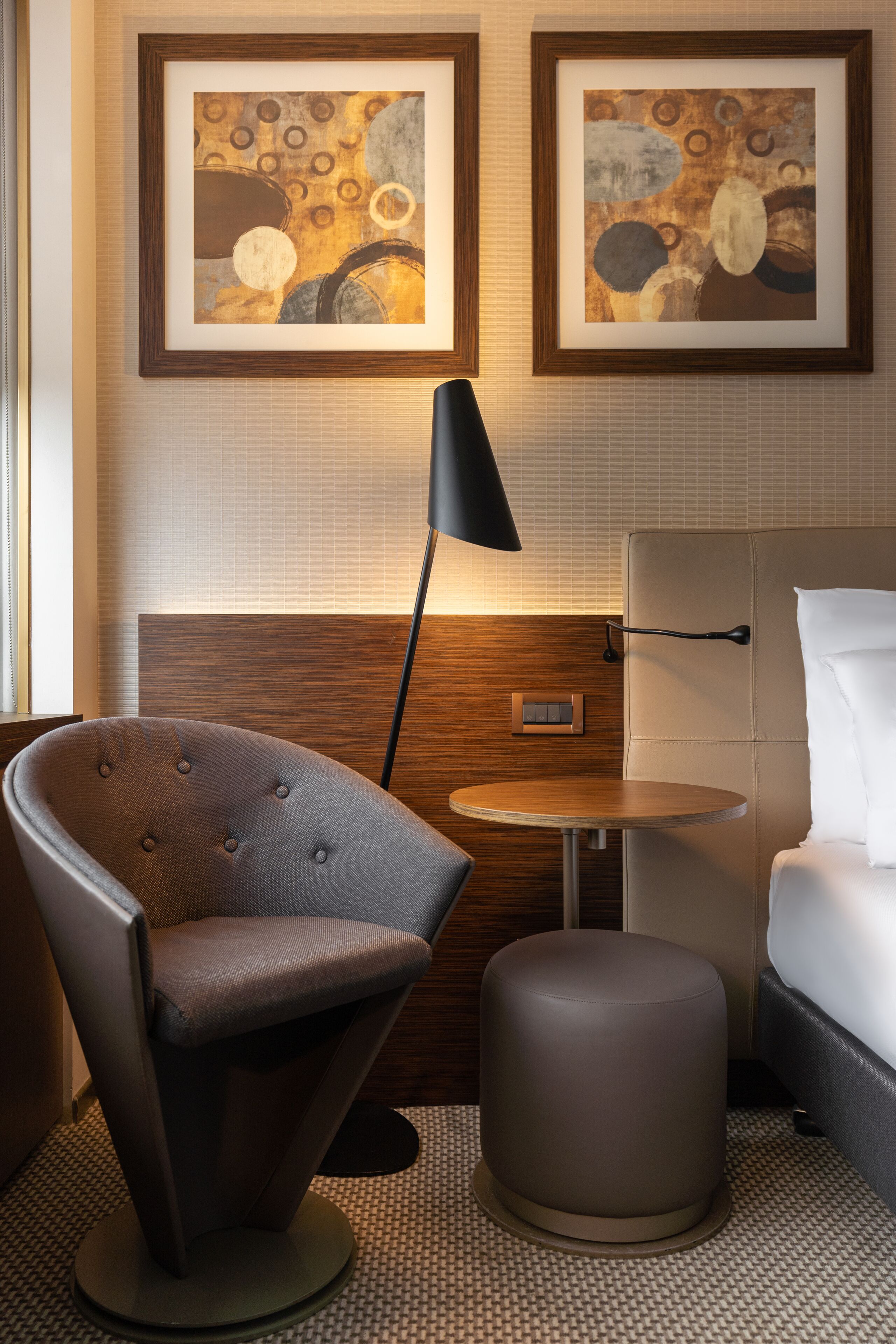 Une chambre d’hôtel avec un fauteuil à capitons, une petite table ronde, une lampe de chevet et deux tableaux abstraits accrochés au mur.