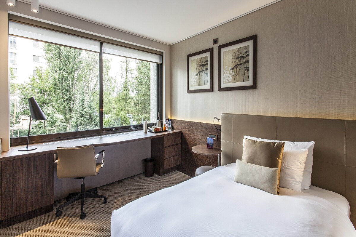 Habitación de hotel con un ventanal grande, un escritorio, una silla, una cama con almohadas y dos obras de arte enmarcadas en la pared.
