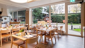 A restaurant with tables and chairs arranged for dining, large windows offering a view of the outdoors, and a visible exit sign. - Crowne Plaza Geneva by IHG (Meyrin)