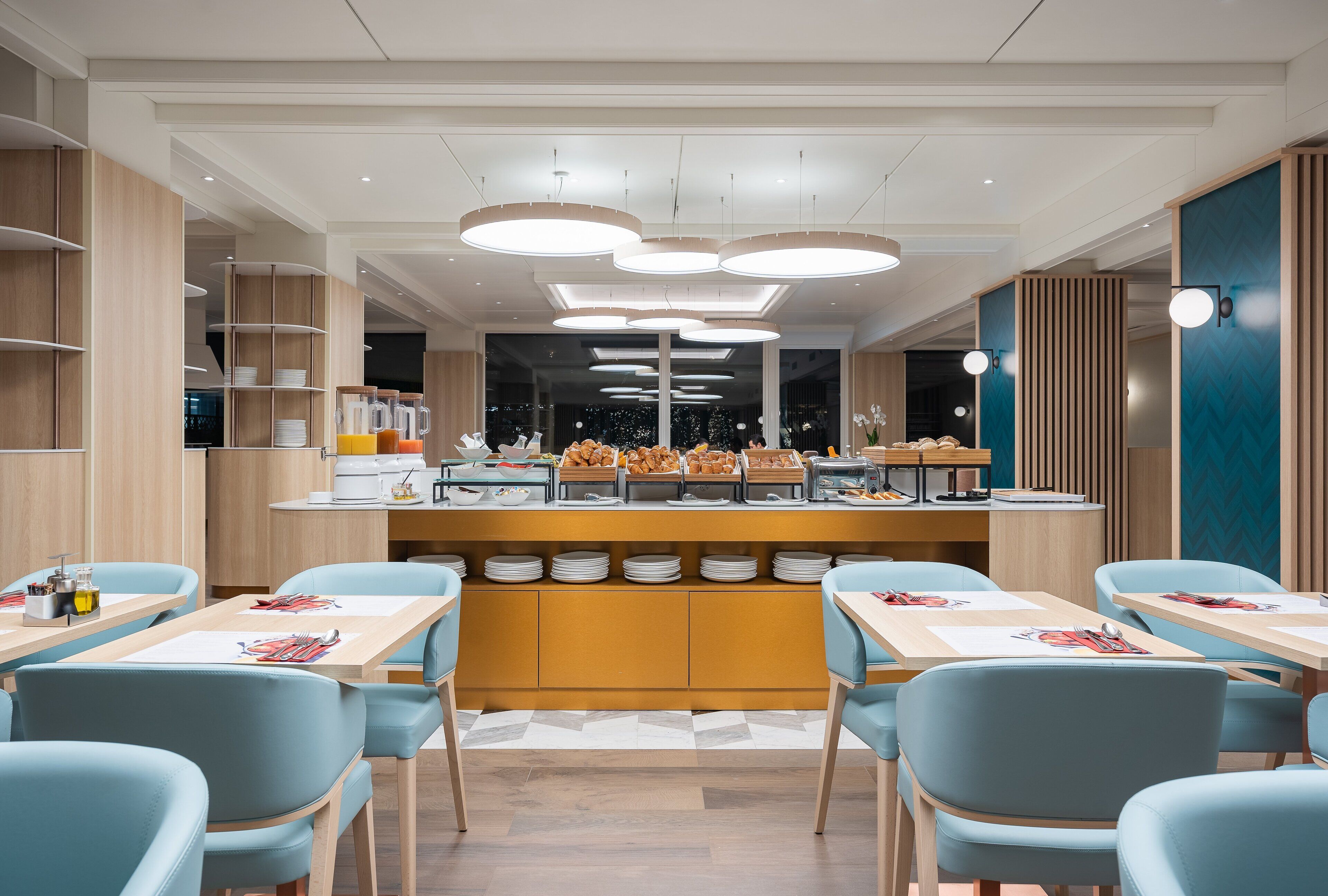 A modern dining area with light blue chairs, a central buffet with various food items, and a wooden counter.