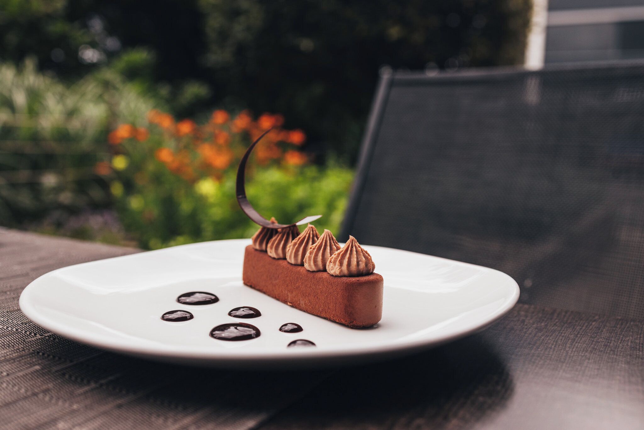 Un dessert servi sur une assiette blanche, décoré de motifs en forme de spirales de chocolat et d’une feuille de chocolat, le tout posé sur une surface texturée.