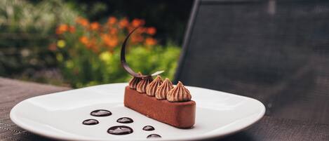 Un dessert servi sur une assiette blanche, décoré de motifs en forme de spirales de chocolat et d’une feuille de chocolat, le tout posé sur une surface texturée.