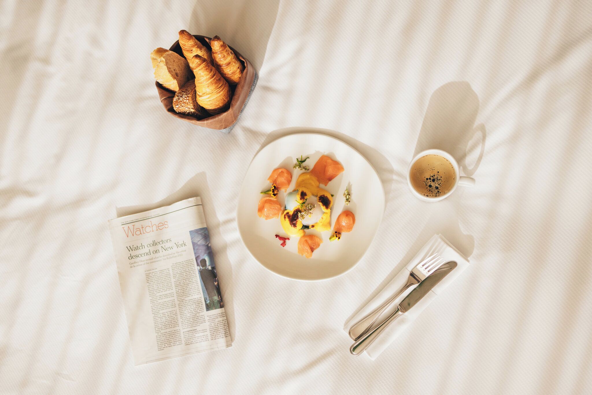 A breakfast setting with croissants, a plate of food, a cup of coffee, and a newspaper on a bed.
