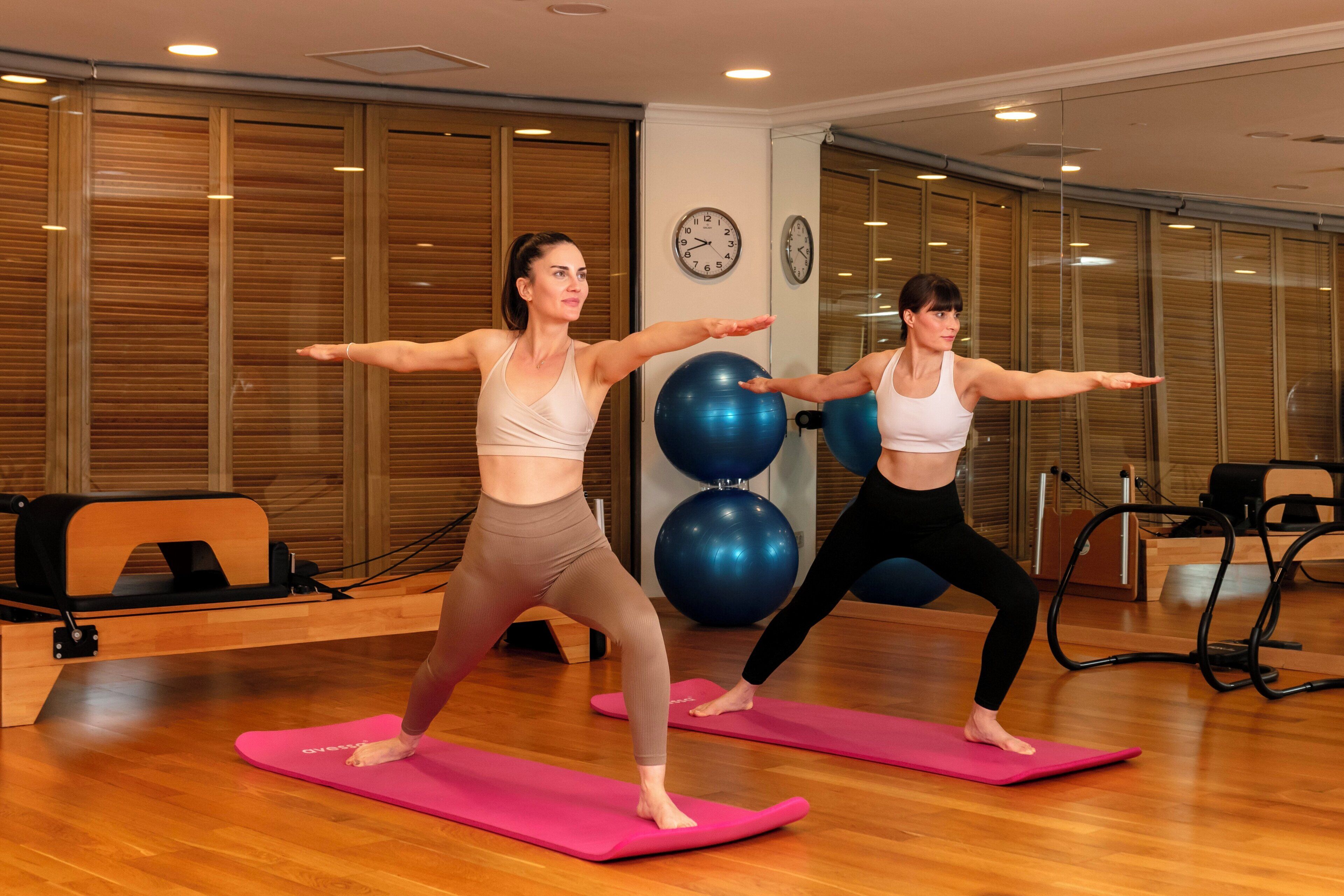 Dos personas practicando yoga sobre colchonetas rosas en una habitación con suelo de madera, pelotas de ejercicio grandes y un reloj en la pared.