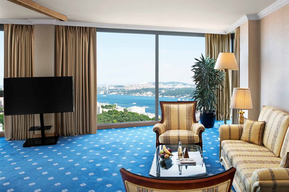 A hotel room with a large window, a television, a sofa, a chair, a coffee table, and a potted plant.