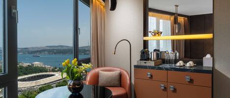 A modern hotel room with a large window offering a city view, a round table with a vase of flowers, a comfortable seating area, and a well-equipped kitchenette.