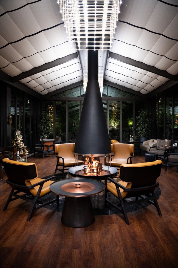 Un salón moderno con chimenea central, asientos de cuero y un techo alto con una lámpara geométrica.