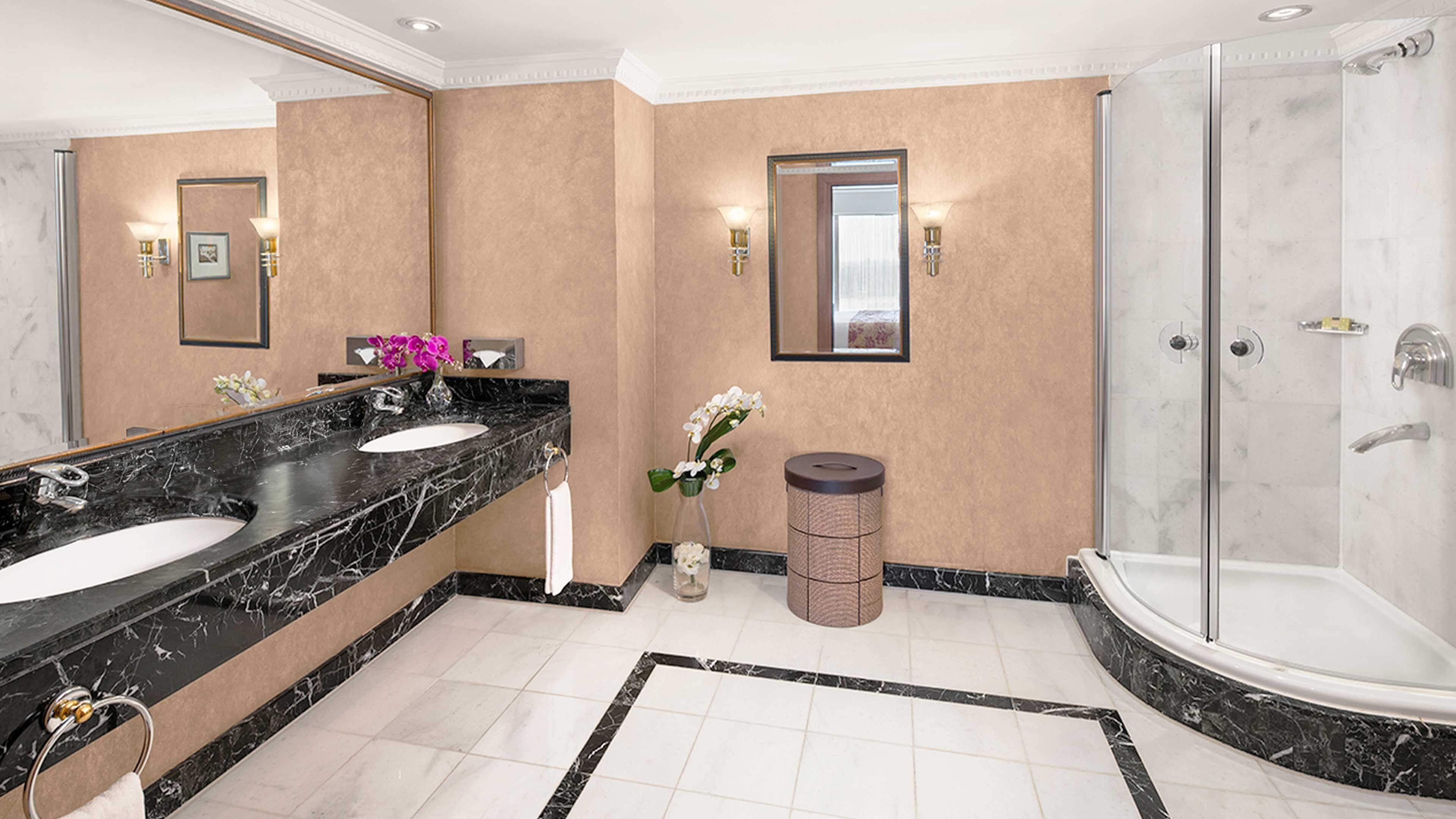 A modern bathroom with two sinks, a large mirror, a shower, and a trash can.