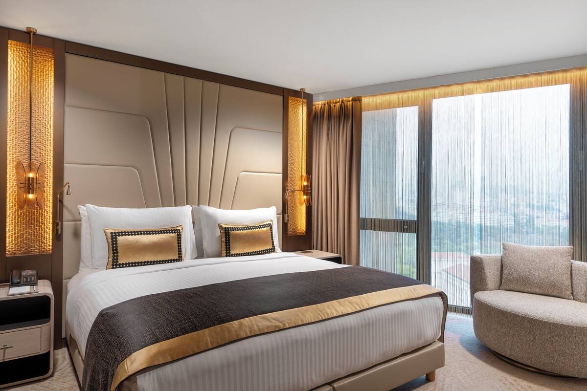 A modern hotel room with a large bed, a sofa, a nightstand with a phone, and a view of the city through large windows.