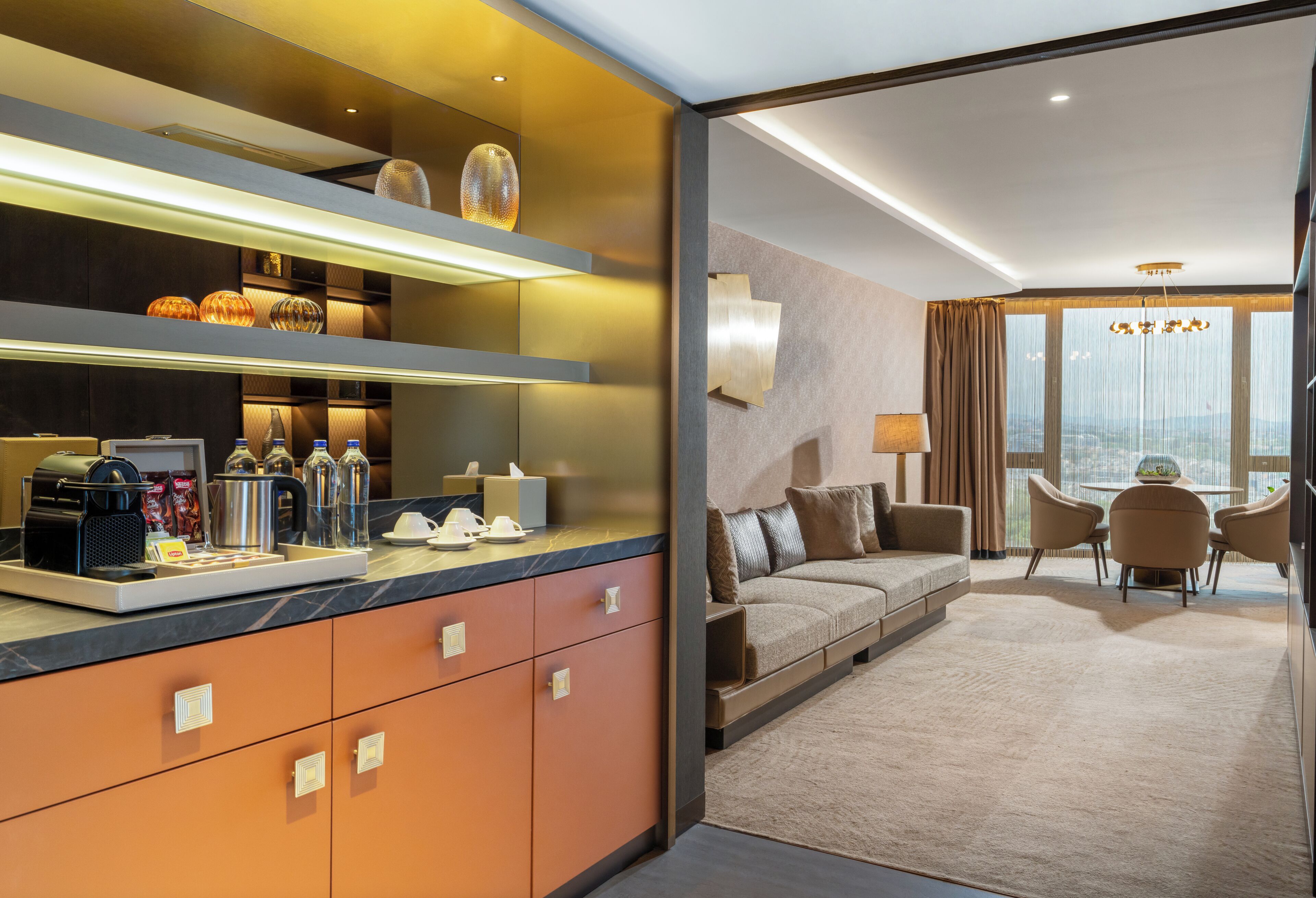 A modern hotel room with a kitchenette, a sofa, a dining area, and a large window offering a city view.