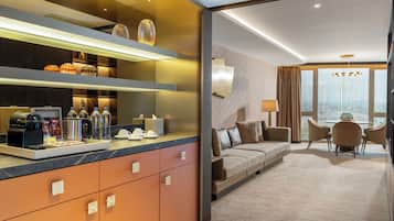 A modern hotel room with a kitchenette, a sofa, a dining area, and a large window offering a city view.