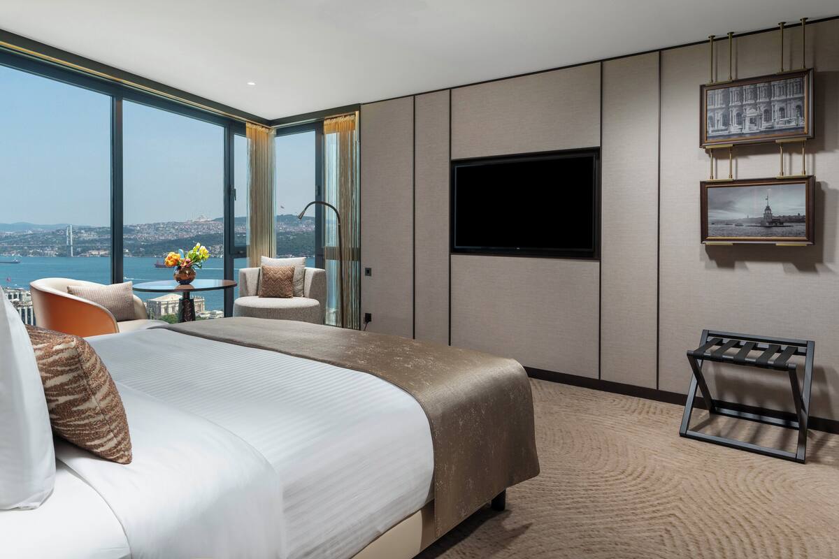 A modern hotel room with a large bed, a TV mounted on the wall, and a view of the city through large windows.
