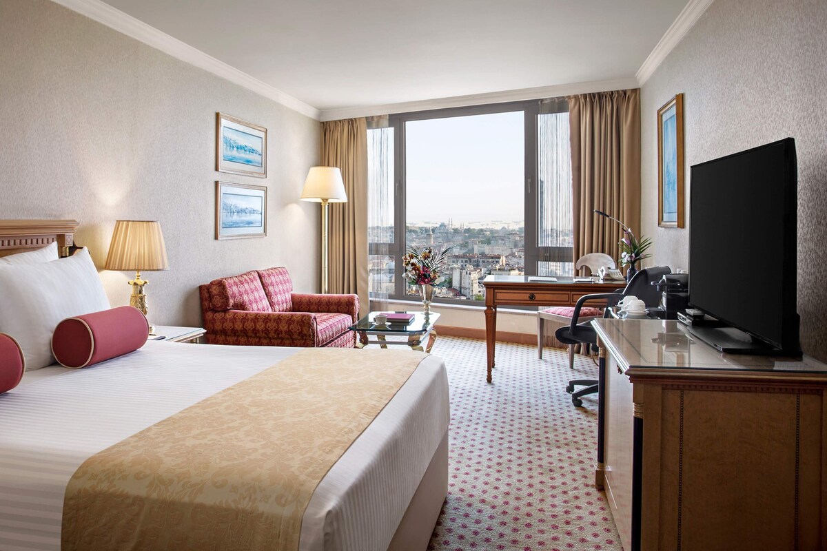 A hotel room with a large bed, a desk, a television, and a view of the city.