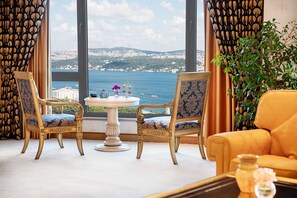 Una habitación con dos sillas ornamentadas y una mesa, vista al mar y edificios distantes, además de una planta grande.