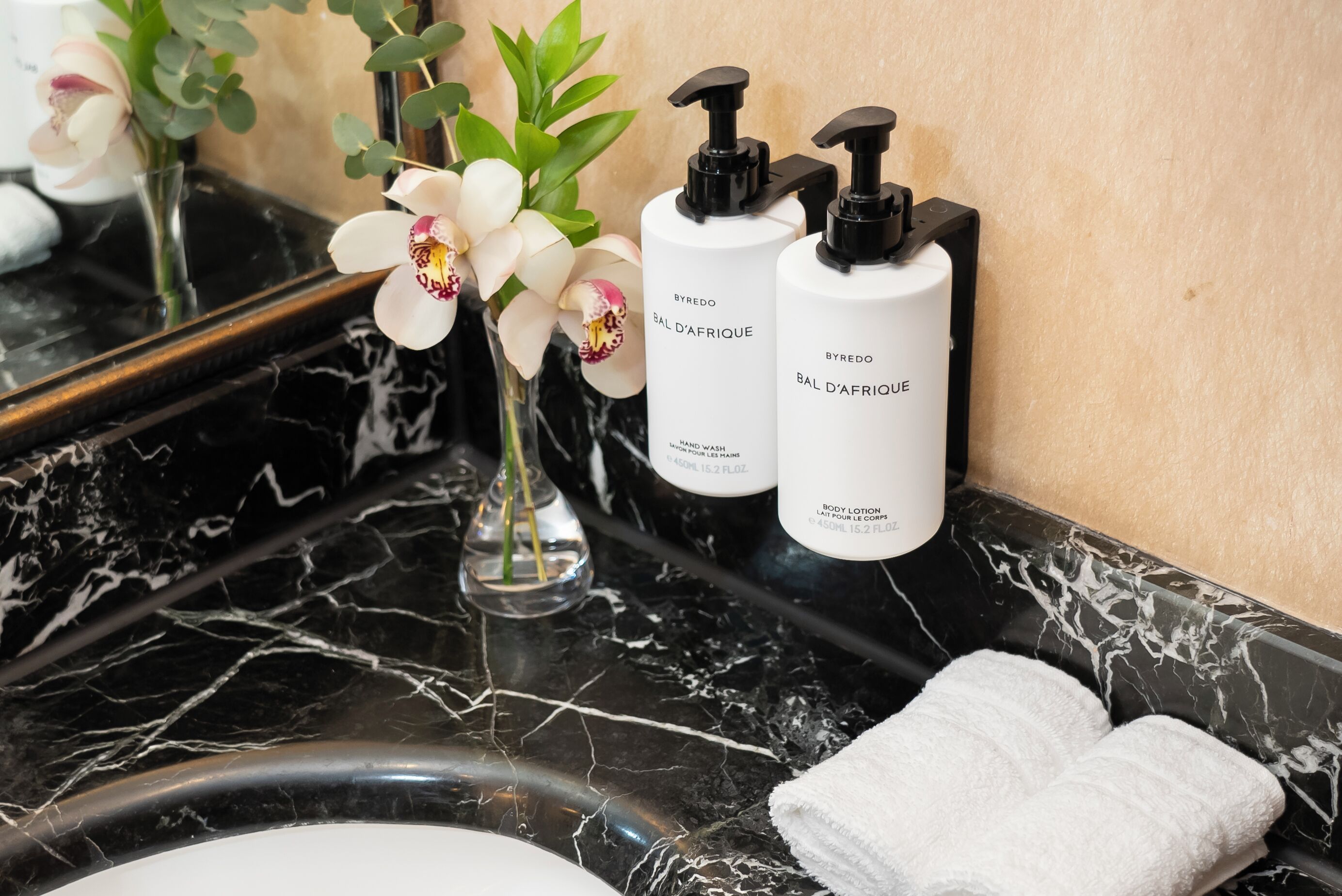 Un tocador de baño con encimera de mármol negro, dos botellas de crema para manos, una toalla blanca y un jarrón con orquídeas blancas.