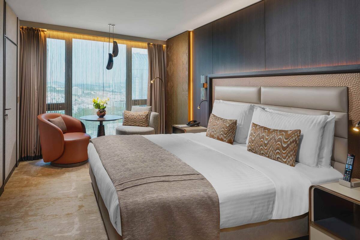 A hotel room with a large bed, a seating area with a chair and a small table, and a TV on the left.